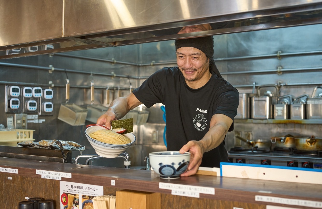 ステンレス製の厨房で、麺を盛った器を持つ黒い服のスタッフ。